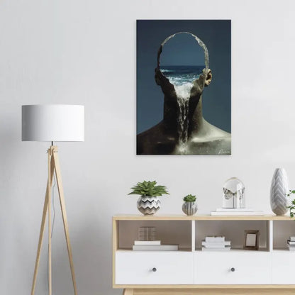Une impression sur toile aux tons sombres représentant une figure surréaliste avec des vagues océaniques s’écoulant à travers sa tête, accrochée sur un mur blanc.
