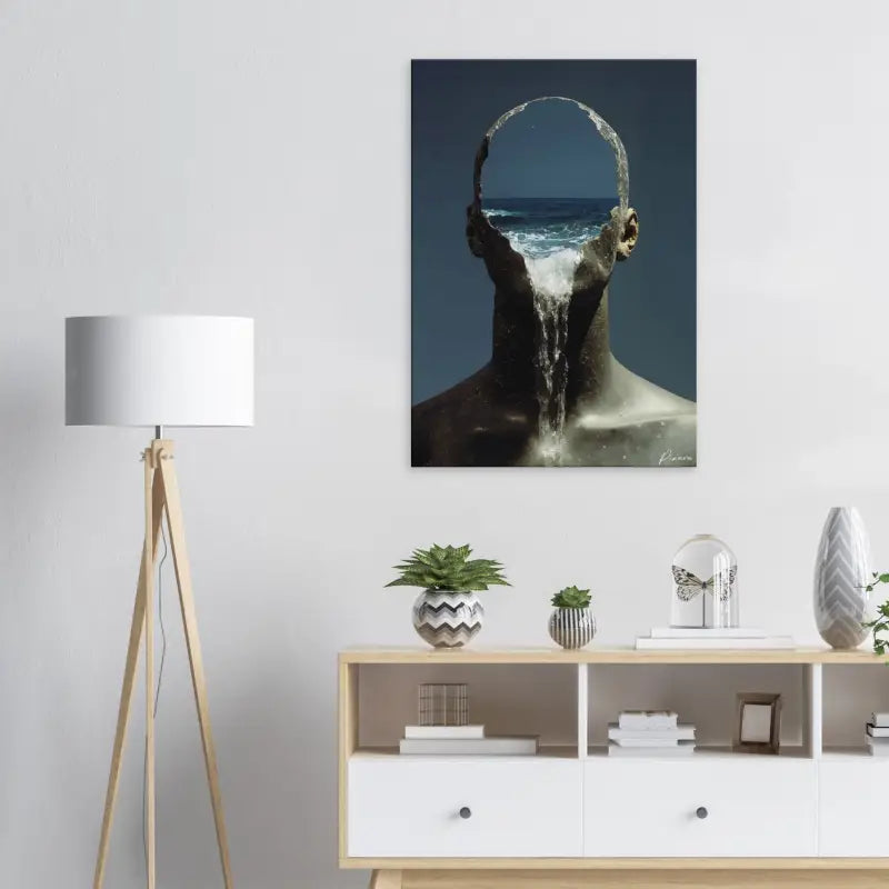 Une impression sur toile aux tons sombres représentant une figure surréaliste avec des vagues océaniques s’écoulant à travers sa tête, accrochée sur un mur blanc.